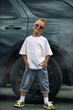 © Vitalii But - Stylish little boy stands confidently in front of a dark SUV wheel, wearing a white oversized t-shirt, denim shorts, and aviator sunglasses, showcasing hip-hop inspired streetwear fashion.