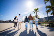© CarlosBarquero - Group of friends walk away along a wide pedestrian promenade in a blue sky bright urban residential district with palm trees and clear blue sky.