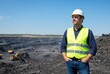 © Latarr - Professional safety-hardhat-clad supervisor overseeing large open-pit coal mine operation with heavy machinery under clear blue sky, confident industrial leadership