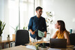 © Studio Marmellata - A young man wearing glasses stands and holds a business document while talking to his female colleague in a modern sunlit office. She listens while at her desk.