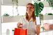 © Studio Marmellata - A smiling woman with red hair holds a vibrant red shopping bag and shows a blank smartphone screen. She is standing in a bright indoor garden area filled with plants.