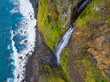 © Amazing Aerial - Aerial view of a waterfall cascading down a steep, mossy, rocky cliff into the Atlantic Ocean, with waves crashing against the dark pebble beach below in Madeira, Portugal.