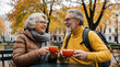 © miss irine - Happy senior couple sits at outdoor cafe table in autumn park. Mature man, woman drink hot coffee, talk, laugh together. Elderly partners enjoy leisure time, companionship, warm weather in city.