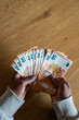 © CarlosBarquero - Vertical. Close-up top view of a Black woman holding and counting a large amount of fifty euro banknotes over a wooden table, representing savings, income, wealth and financial planning.