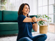 © ONE-STUDIO - Cheerful middle-aged woman stretching her arm, sitting on a rug at home, promoting wellness.
