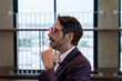 © Windscale - A white man with glasses in a stylish wine-colored suit sitting on a red chair in a well-lit room with windows in the background