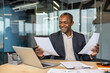 © Liubomir - Smiling businessman reviewing documents, working at an office desk with a laptop and various paperwork, representing success and diligent professional work