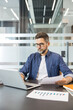 © Liubomir - Professional man wearing glasses working with a laptop and holding documents, focusing on business analysis in a modern office environment