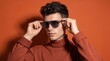 © Yassine - Handsome young man wearing stylish grey sunglasses and a cozy brown turtleneck sweater posing against a vibrant solid orange background
