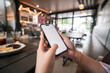 © faizal - Person holds phone with blank screen, fingers gently gripping edges. Restaurant background softly blurred, warm lights glow overhead. Fingers show casual, relaxed grip, suggesting leisurely use