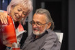 © Anelo - Smiling elderly woman pouring drink for seated man at home