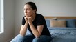 © NimVisuals - A thoughtful middle-aged woman rests her chin on her hands while sitting on a bed.