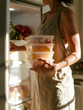 © lemonmoon - Woman in linen apron holding clear food container stack beside open refrigerator in sunlit kitchen with fresh groceries and cropped face
