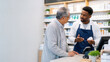© SerPak - Pharmacist in an apron consults a senior man at a pharmacy counter, holding a pill bottle while shelves of medications fill the background.