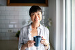 © Marko Geber - Smiling woman holding coffee mug at home kitchen window