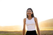 © liliyabatyrova - In a serene setting filled with warm hues, a young girl stands with a radiant smile as the sun sets behind rolling hills. Her carefree posture captures the beauty of nature