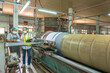 © BESTIMAGE - Textile inspection factory teamwork female supervisor in safety vest checking and discussing yarn production quality with woman worker in textile machine mill holding thread spool and clipboard.