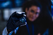 © ultramansk - Gloved hand presenting a microprocessor chip close to camera in a dark blue-lit cybersecurity lab with security dashboards glowing in the blurred background.