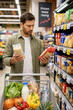 © elenakibrik - Concerned Man Comparing Food Prices in Supermarket Amid Rising Cost of Living