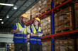 © ultramansk - Male warehouse supervisor with a beard and his female coworker in protective workwear hold a two way radio while checking the racks in a large logistics distribution centre with tall.