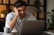 © Anastasiia - A man with dark hair is sitting at a desk, looking stressed while resting his head on his hand and staring at an open laptop in an indoor setting.