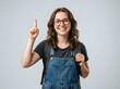 © GraphixStudio - Confident young woman in denim overalls and glasses pointing upward with a cheerful smile against neutral background