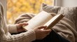 © IvanMasyudi - Cozy autumn reading by window, woman holding open book with warm blanket, peaceful fall lifestyle scene with yellow foliage