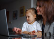 © Seleznov_Molchanova - mother with a child sits at a table and works on a laptop. remote work mom baby concept. Work-from-home mom focused on her laptop, managing online tasks while balancing motherhood and career.