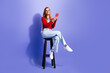 © deagreez - Young woman sitting on a stool smiling while using a smartphone against a purple studio background in casual jeans and red top