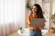 © Prostock-studio - Smiling Young Latin American Woman Holding Laptop While Standing In Bright Home Office, Showing Happiness And Confidence. Engaged In Work Or Online Communication, Modern Business Concept.