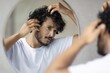 © Prostock-studio - Focused Young Man With Curly Hair Inspecting Hair In Mirror At Home, Taking Care Of Appearance And Grooming Routine, Engaged In Self-care And Personal Care At Home.