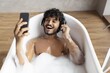 © Prostock-studio - Smiling Young Man With Curly Hair, Enjoying Bathing In Bathtub With Foam, Wearing Headphones, Using Smartphone In Modern Bathroom, Relaxing And Unwinding At Home Spa Setting.