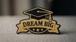 © Panuwat - Embroidered patch with a graduation cap and gold Dream Big phrase on dark fabric.