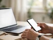 © aziseptian - Hands holding smartphone with blank screen next to laptop on desk