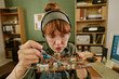 © AnnaStills - Young adult Caucasian woman wearing safety glasses repairing vintage circuit board in retro office. Image suitable for engineering education and electronics repair service marketing
