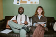 © AnnaStills - Portrait of diverse young adult colleagues sitting on sofa in retro office, holding coffee cup and documents. Image represents vintage corporate culture, suitable for HR marketing