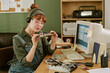 © AnnaStills - Young adult Caucasian woman wearing safety glasses and headphones examining circuit board at retro office desk. Image represents vintage engineering and hardware repair services