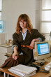 © AnnaStills - Portrait of Caucasian young adult woman sitting on desk in retro office next to vintage computer monitor, representing corporate nostalgia and 1990s workplace culture, vertical