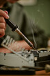 © AnnaStills - Vertical image of hand of Caucasian young adult holding soldering iron over vintage electronic device with smoke rising, representing retro office repair and engineering concepts