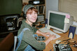 © AnnaStills - Portrait of smiling young adult Caucasian woman wearing headset, typing on retro computer keyboard in vintage office, representing nostalgia marketing and tech history
