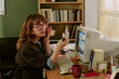 © AnnaStills - Young adult Caucasian woman looking in small mirror at desk in retro office, adjusting makeup near vintage computer. Image suitable for nostalgic corporate marketing campaigns