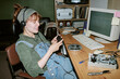 © AnnaStills - Young adult Caucasian woman wearing headphones and holding old pager in retro office, repairing vintage computer hardware. Image suitable for nostalgic tech marketing campaigns