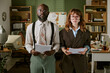 © AnnaStills - Portrait of African American middle aged man and Caucasian young woman standing in retro office holding documents. Image represents vintage corporate culture and diverse teamwork