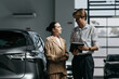 © wedmoments.stock - Woman consulting with a salesperson beside a new car in a modern automotive showroom. Concept car shopping, customer service and vehicle selection.