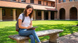 © Celt Studio - Smiling young female college student wearing casual clothes sitting outside upon stone bench using digital tablet with stylus pen near classic brick campus buildings
