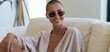 © ckybe - Smiling woman with short hair lounges on a sleek sofa, wearing casual attire and trendy sunglasses in a bright, minimalist space.
