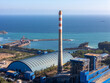 © Amazing Aerial - Aerial view of a power plant with a tall striped chimney, storage warehouse, and industrial facilities along the coastline with a cargo ship in the harbor in Pacitan, East Java, Indonesia.