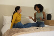 © WavebreakMediaMicro - Diverse female friends sitting on bed talking in room, mustard and striped tops with smartphone