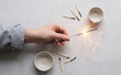 © SimpleDesignStudio - Person holding a burning sparkler with matches and small ceramic bowls on a light textured background