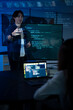 © Velocitystock777 - Smiling Asian male software developer gesturing during a presentation with programming code projected on a screen in a blue-lit room